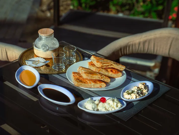 A breakfast tray with pastries, cheese dishes, honey and glasses of milk on an outdoor table in morning light.