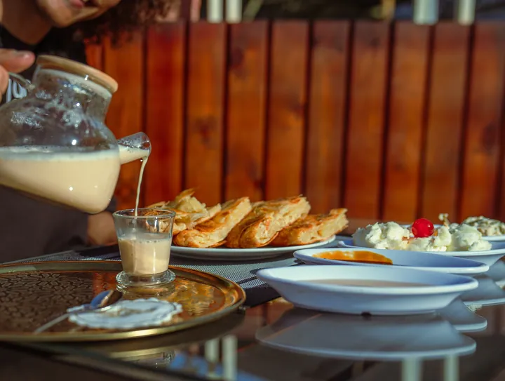 A hand pouring a milky drink from a glass jug into a cup beside plates of pastries and mezze.