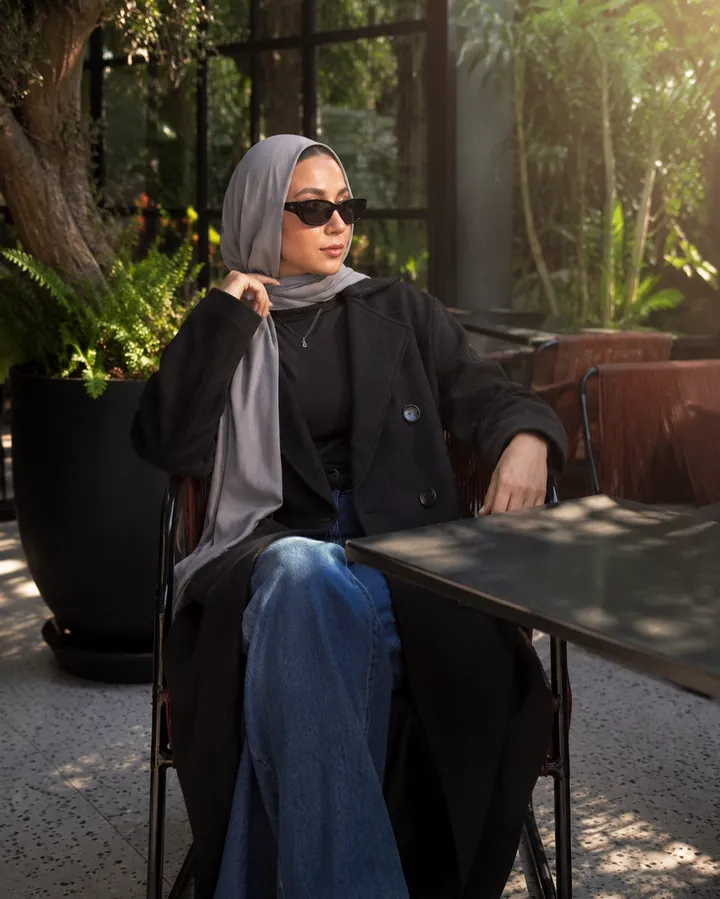 Seated at an outdoor café table in a black coat, wide-leg jeans and a grey headscarf.
