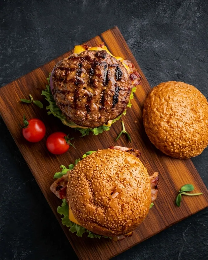 Overhead frame of two grilled burgers on a wooden board with sesame buns and tomatoes.