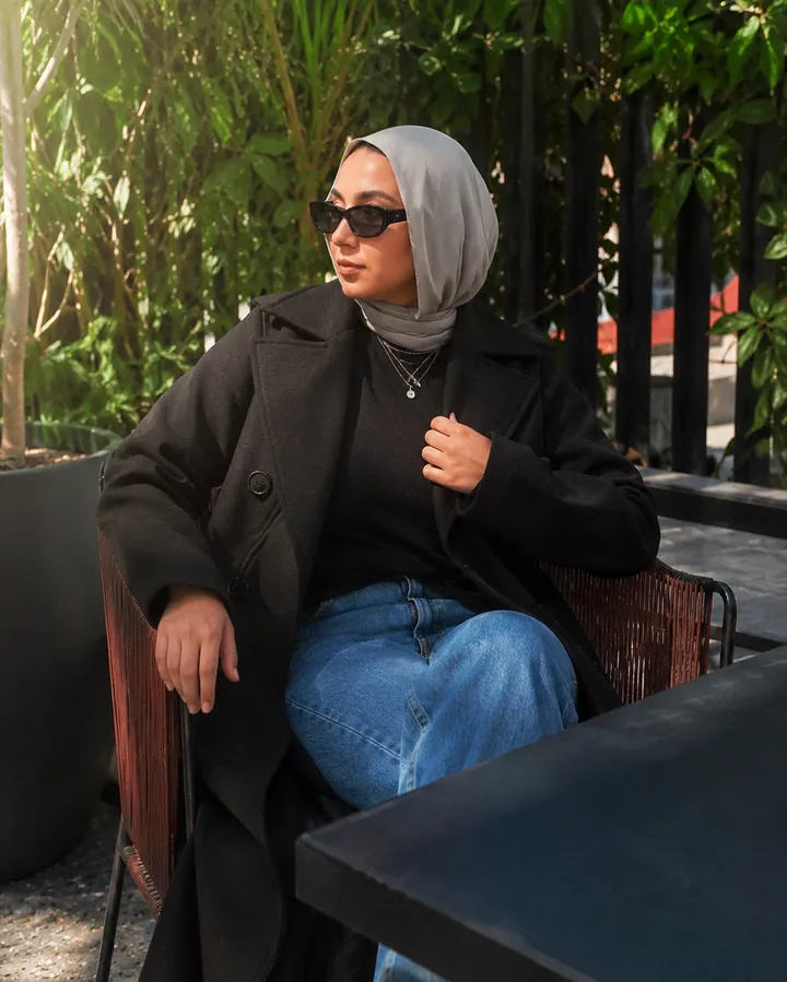 A woman in a long black coat and grey headscarf with sunglasses, seated on a plant-filled café terrace.