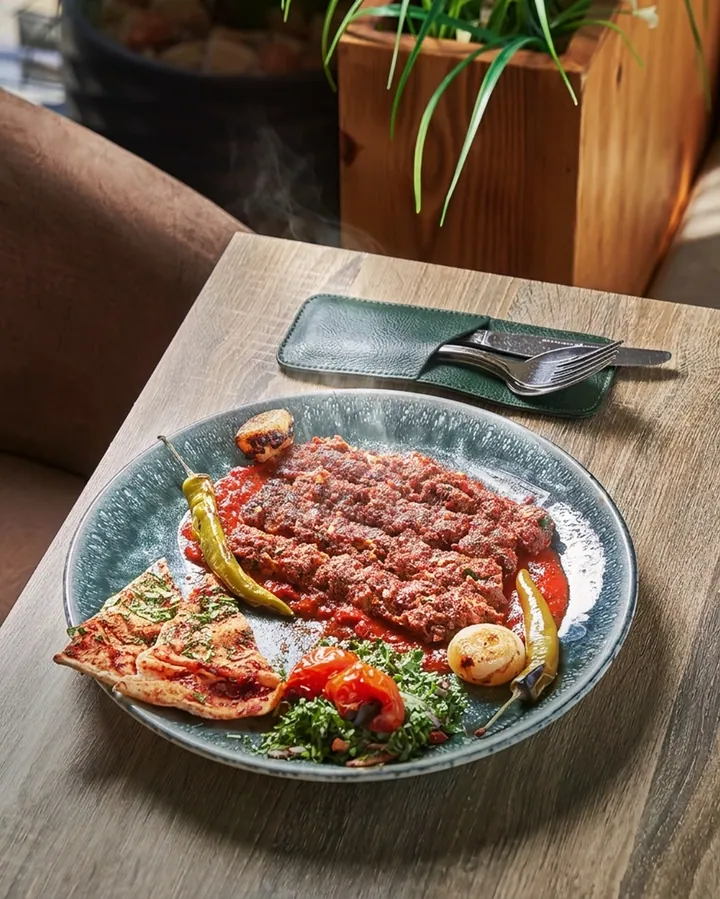 A kofta plate with grilled tomato, chilli and flatbread, steam rising, on a wooden table.