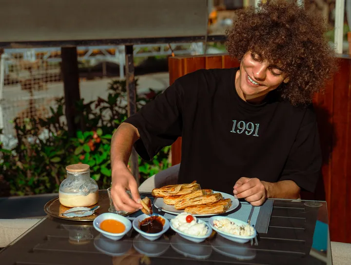 A smiling young man serving himself a piece of pastry from an open-air breakfast spread.