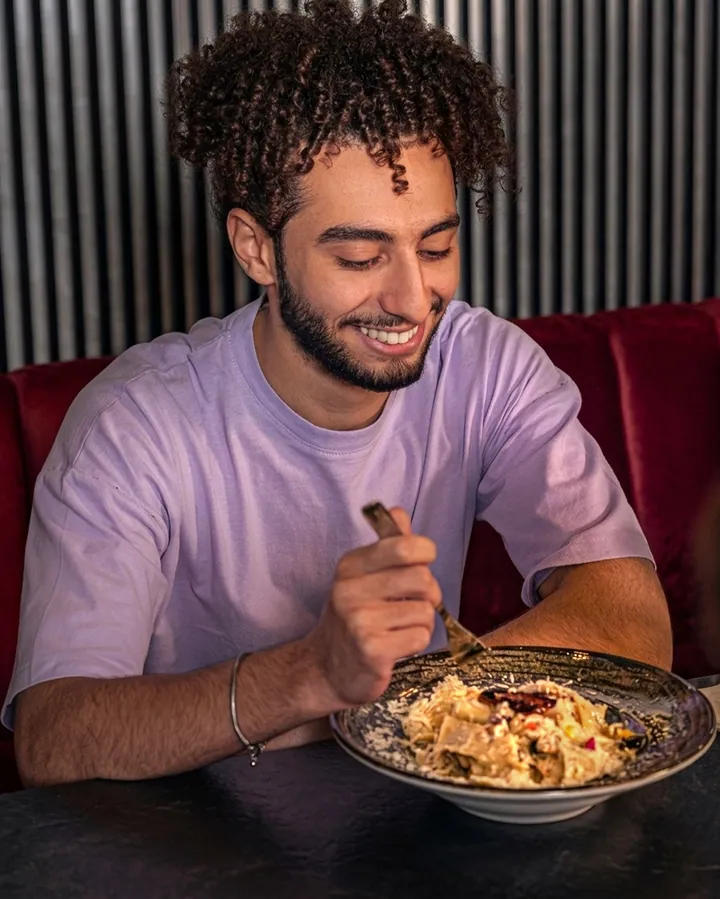 A young man eating pasta from a dark bowl inside a warmly lit restaurant.