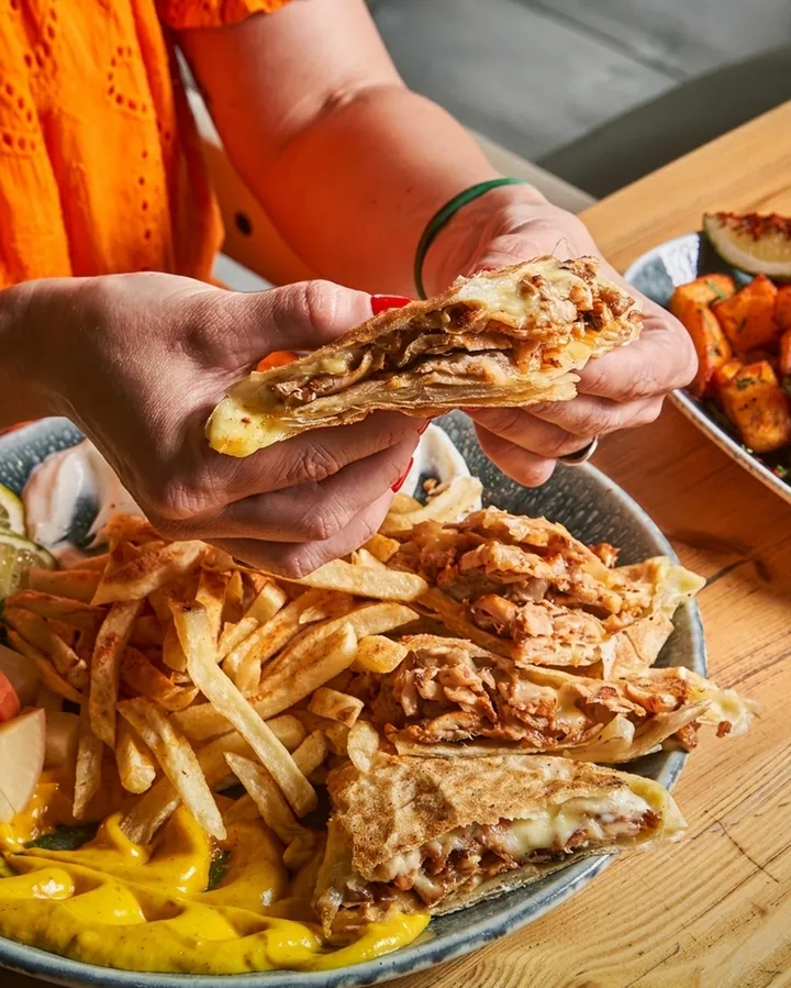A hand holding a cheese-filled quesadilla wedge above a large platter of fries and wraps.